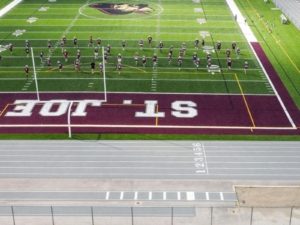 A high-angle view of a football practice session at a synthetic turf field marked “ST. JOE’S” in large white letters on a maroon end zone. Players in maroon and white uniforms are running drills on the field, with a large team logo at midfield. In the foreground, a gray running track with clearly marked lanes (1–6) wraps around the field, and a goalpost frames the scene near the bottom of the image.