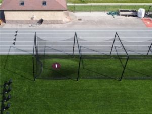 An aerial view of a batting cage setup on a green artificial turf field, enclosed by black netting supported by tall poles. In the background, a gray running track with six lanes and black arrow markings runs parallel to a tan utility building with a reddish-brown roof. Additional athletic equipment and storage materials are visible behind the track near a fenced area.